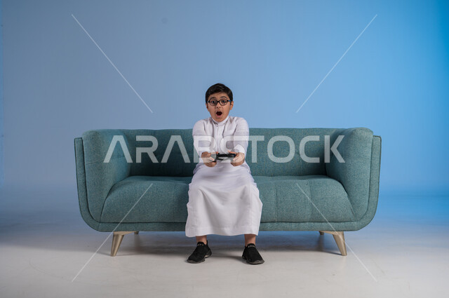 ايماءات الاندماج والذهول والصدمة، استخدام الأجهزة والتكنولوجية والتقنيات الحديثة، بورتريه لفتى عربي خليجي سعودي يرتدي الثوب التقليدي والنظارة الطبية يجلس على الكنبة ويمسك بيده عصا التحكم، اللعب بألعاب الفيديو والبلايستيشين الترفيهية، خلفية زرقاء