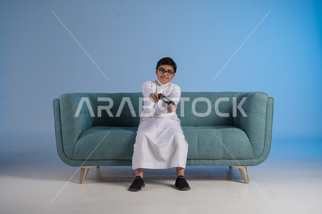 Having a fun and entertaining time playing, interacting and merging with PlayStation games, modern entertainment electronic technologies, portrait of a Saudi Arabian Gulf boy wearing traditional dress and glasses sitting on a comfortable chair holding a joystick in his hand playing video games, blue background