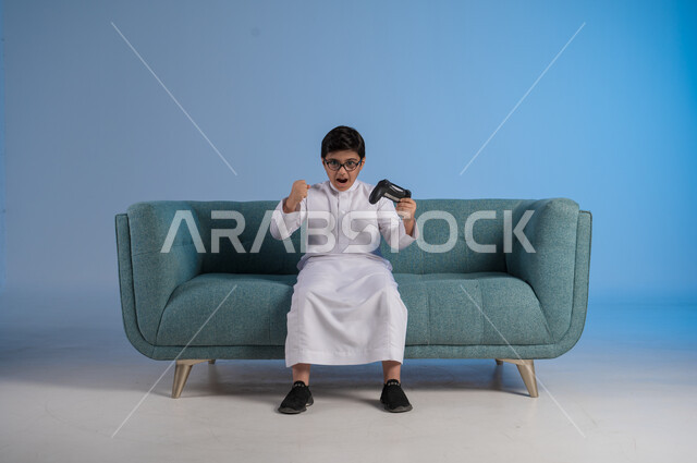 قضاء وقت ممتع ومسلي باللعب، التفاعل والاندماج مع ألعاب البلايستيشن، تقنيات الكترونية حديثة ترفيهيه, بورتريه لفتى عربي سعودي خليجي يرتدي الثوب التقليدي والنظارة الطبية يجلس على كرسي مريح يحمل بيده عصا التحكم يقوم باللعب في ألعاب الفيديو، خلفية زرقاء