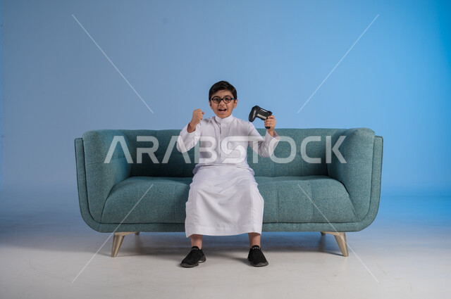 Enjoying playing entertaining electronic games, using technological devices and modern techniques, portrait of a Saudi Arabian Gulf boy wearing traditional dress and glasses sitting on a chair holding a joystick with gestures of encouragement and enthusiasm, blue background
