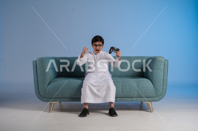 Enjoying playing entertaining electronic games, using technological devices and modern techniques, portrait of a Saudi Arabian Gulf boy wearing traditional dress and glasses sitting on a chair holding a joystick with gestures of encouragement and enthusiasm, blue background
