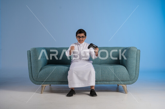 الأنشطة الترفيهية الممتعة, التقنيات الحديثة والتكنولوجيا المتطورة, قضاء أوقات الفراغ بألعاب البلايستيشن, بورتريه لفتى عربي خليجي سعودي يرتدي الثوب التقليدي والنظارة الطبية يمسك بيده عصا التحكم بتفاعل واندماج لتحقيق الفوز والانتصار, خلفية زرقاء