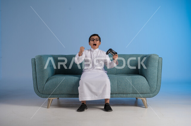 تحقيق الفوز والانتصار, الأنشطة الترفيهية الممتعة, التقنيات الحديثة والتكنولوجيا المتطورة, قضاء أوقات الفراغ بألعاب البلايستيشن, بورتريه لفتى عربي خليجي سعودي يرتدي الثوب التقليدي والنظارة الطبية يمسك بيده عصا التحكم بتفاعل واندماج, خلفية زرقاء