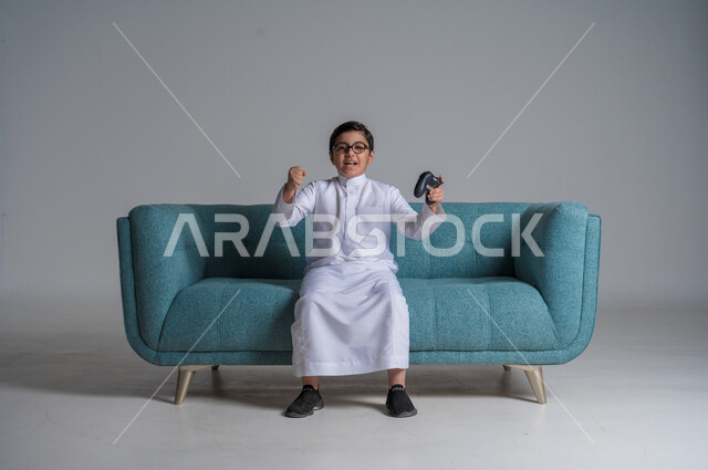 Spending free time playing PlayStation games, portrait of a Saudi Arabian Gulf boy wearing traditional dress and glasses holding a joystick in his hand interacting and merging, joy and happiness for achieving victory, fun recreational activities, modern technologies and advanced technology, blue background