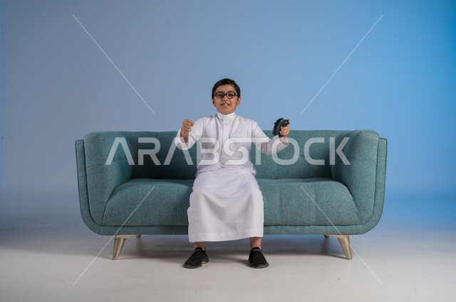 الفرح والسعادة لتحقيق الفوز, الأنشطة الترفيهية الممتعة, التقنيات الحديثة والتكنولوجيا المتطورة, قضاء أوقات الفراغ بألعاب البلايستيشن, بورتريه لفتى عربي خليجي سعودي يرتدي الثوب التقليدي والنظارة الطبية يمسك بيده عصا التحكم بتفاعل واندماج, خلفية زرقاء