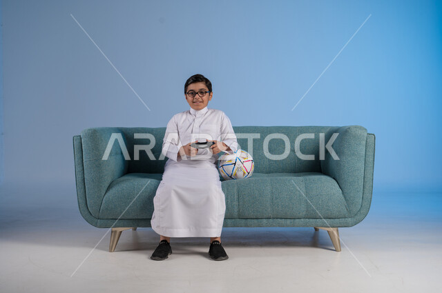  التقنيات الحديثة والتكنولوجيا المتطورة, قضاء أوقات الفراغ بألعاب البلايستيشن, بورتريه لفتى عربي خليجي سعودي يرتدي الثوب التقليدي والنظارة الطبية يمسك بيده عصا التحكم بتفاعل واندماج لتحقيق الفوز والانتصار, الأنشطة الترفيهية الممتعة, خلفية زرقاء