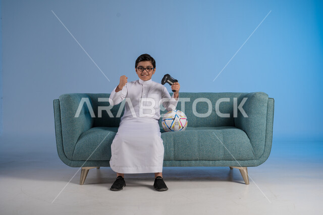 الفرح والسعادة لتحقيق الفوز, الأنشطة الترفيهية الممتعة, التقنيات الحديثة والتكنولوجيا المتطورة, قضاء أوقات الفراغ بألعاب البلايستيشن, بورتريه لفتى عربي خليجي سعودي يرتدي الثوب التقليدي والنظارة الطبية يمسك بيده عصا التحكم بتفاعل واندماج, خلفية زرقاء