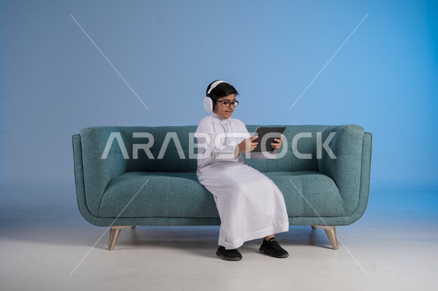 تقنيات وتكنولوجية متطورة, القيام بالبث المباشر عبر وسائل التواصل الاجتماعي, إجراء مكالمة فيديو عبر التابليت, بورتريه لفتى عربي سعودي خليجي مبتسم يرتدي الثوب التقليدي والنظارة الطبية يجلس على كنبه مريحة يقوم بالتقاط صور سيلفي تذكارية, خلفية زرقاء