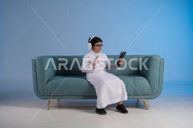 Live streaming on social media, making a video call via tablet, portrait of a smiling Saudi Arabian Gulf boy wearing traditional thobe and glasses sitting on a comfortable sofa holding a tablet, advanced techniques and technology, blue background