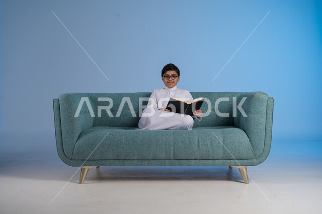 حب المطالعة والقراءة, قضاء أوقات الفراغ بالتثقيف والتعلم, تنمية مهارات القراءة, الاهتمام بتصفح الكتب المفيدة, بورتريه لفتى عربي خليجي سعودي يرتدي الثوب التقليدي والنظارة الطبية يجلس على كنبه مريحة ويمسك بيده الكتاب, خلفية زرقاء