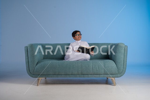 التمعن والنظر إلى شيء ما, محاولة تذكر أمر معين, حب المطالعة والقراءة, قضاء أوقات الفراغ بالتثقيف والتعلم, تنمية مهارات القراءة, بورتريه لفتى عربي خليجي سعودي يرتدي الثوب التقليدي والنظارة الطبية يجلس على كنبه مريحة ويمسك بيده الكتاب, خلفية زرقاء