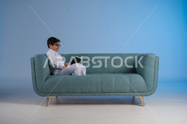 التركيز والاندماج بالمطالعة والقراءة, قضاء أوقات الفراغ بالتثقيف والتعلم, تنمية مهارات القراءة, الاهتمام بتصفح الكتب المفيدة, بورتريه لفتى عربي خليجي سعودي يرتدي الثوب التقليدي والنظارة الطبية يجلس على كنبه مريحة ويمسك بيده الكتاب, خلفية زرقاء