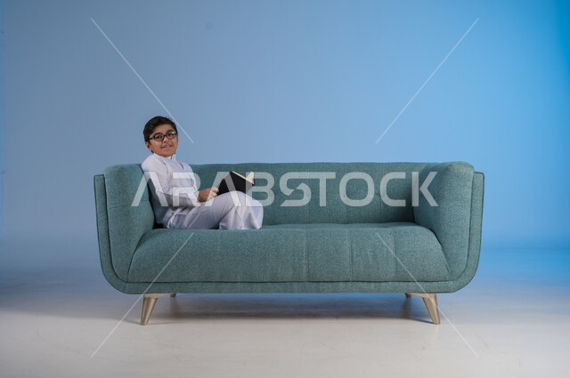 تنمية مهارات القراءة, الاهتمام بتصفح الكتب المفيدة, بورتريه لفتى عربي خليجي سعودي يرتدي الثوب التقليدي والنظارة الطبية يجلس على كنبه مريحة ويمسك بيده الكتاب, حب المطالعة والقراءة, قضاء أوقات الفراغ بالتثقيف والتعلم, خلفية زرقاء