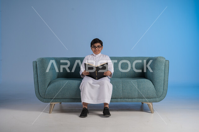 قضاء أوقات الفراغ بالتثقيف والتعلم, تنمية مهارات القراءة, الاهتمام بتصفح الكتب المفيدة, بورتريه لفتى عربي خليجي سعودي يرتدي الثوب التقليدي والنظارة الطبية يجلس على كنبه مريحة ويمسك بيده الكتاب, حب المطالعة والقراءة, خلفية زرقاء