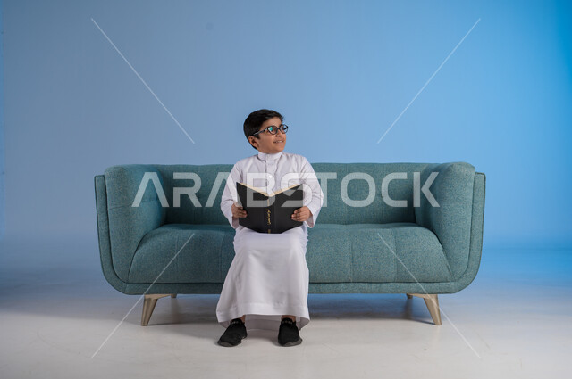 النظر إلى شيء ما بتمعن وتركيز, حب المطالعة والقراءة, قضاء أوقات الفراغ بالتثقيف والتعلم, تنمية مهارات القراءة, الاهتمام بتصفح الكتب المفيدة, بورتريه لفتى عربي خليجي سعودي يرتدي الثوب التقليدي والنظارة الطبية يجلس على كنبه مريحة ويمسك بيده الكتاب, خلفية زرقاء