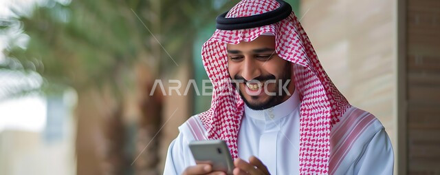 Chatting with friends via mobile programs and applications, browsing social networking sites, a Saudi Arabian Gulf man wearing a traditional shemagh and thobe holding a mobile phone in his hand, integrating modern technologies and technology into practical life, gestures of happiness and pleasure when hearing good news