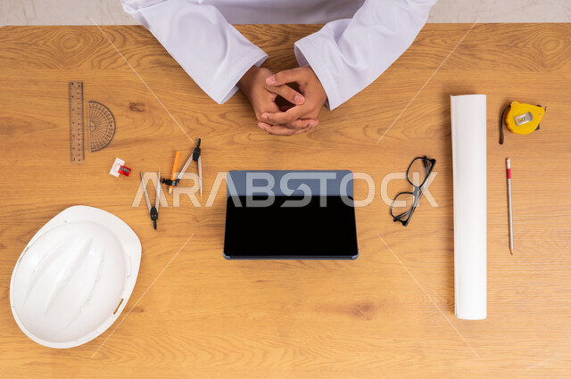 التسوق عبر المواقع الالكترونية، صورة علوية عامودية ليدين رجل عربي خليجي سعودي يستخدم جهاز التابليت وبجانبه خوذة الحماية والأدوات الهندسية، عرض شاشة سوداء فارغة، الحصول على المستلزمات اللازمة اونلاين، مفهوم الهندسة والإنشاءات المعمارية، دمج التقنيات الحديثة بالعمل