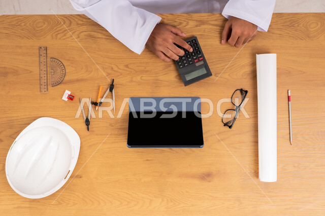 Concept of engineering and architectural construction, integration of modern technologies in work, vertical top view of the hands of a Saudi Arabian Gulf man using a calculator next to a protective helmet and engineering tools, displaying a blank black screen on a tablet, shopping and obtaining the necessary supplies online