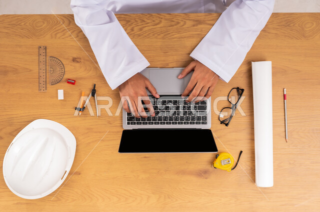 الحصول على المستلزمات اللازمة اونلاين، مفهوم الهندسة والإنشاءات المعمارية، دمج التقنيات الحديثة بالعمل، التسوق عبر المواقع الالكترونية، صورة علوية عامودية ليدين رجل عربي خليجي سعودي يستخدم جهاز اللابتوب وبجانبه خوذة الحماية والأدوات الهندسية، عرض شاشة سوداء فارغة