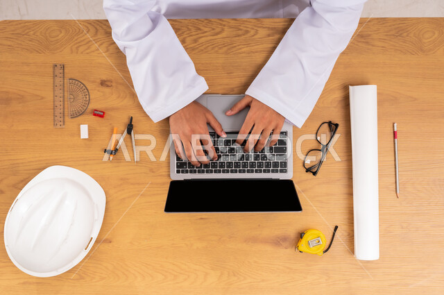 عرض شاشة سوداء فارغة، الحصول على المستلزمات اللازمة اونلاين، مفهوم الهندسة والإنشاءات المعمارية، دمج التقنيات الحديثة بالعمل، التسوق عبر المواقع الالكترونية، صورة علوية عامودية ليدين رجل عربي خليجي سعودي يستخدم جهاز اللابتوب وبجانبه خوذة الحماية والأدوات الهندسية