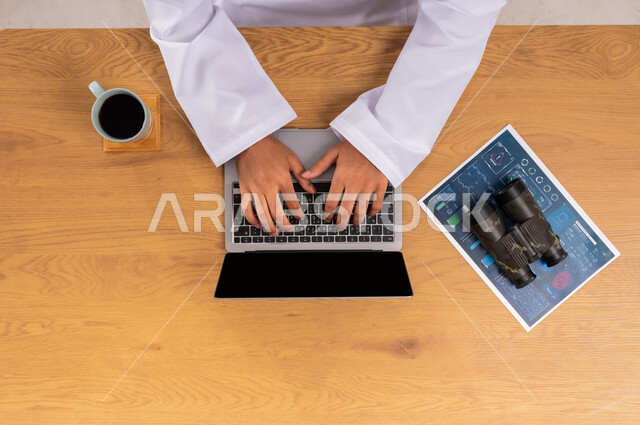 استخدام التقنيات الحديثة في مجال الأمن، البحث لكشف حقائق الجرائم، عرض شاشة سوداء فارغة، صورة علوية عامودية لرجل عربي سعودي خليجي يعمل على جهاز الحاسب المحمول بجانبه منظار وورقة المعلومات والأدلة، مفهوم مسح البصمات، التطور الالكتروني والنمو التكنولوجي