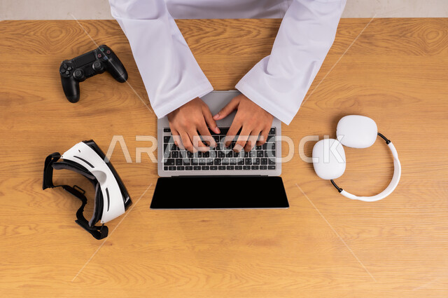 الاستفادة من التكنولوجيا الحديثة، ممارسة الهوايات المختلفة في وقت الفراغ، عرض شاشة سوداء فارغة، صورة علوية عامودية ليدين رجل عربي خليجي سعودي يستخدم جهاز اللابتوب وبجانبه الهيدفون ونظارة الفي آر وقبضة التحكم، مفهوم الإصغاء والاستماع، محاكاة العالم الافتراضي