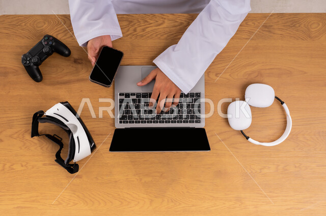 ممارسة الهوايات المختلفة في وقت الفراغ، عرض شاشة سوداء فارغة، صورة علوية عامودية ليدين رجل عربي خليجي سعودي يستخدم اللابتوب والجوال بجانبه الهيدفون ونظارة الفي آر وقبضة التحكم، مفهوم الإصغاء والاستماع، محاكاة العالم الافتراضي، الاستفادة من التكنولوجيا الحديثة