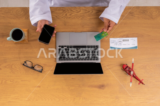 الدفع بواسطة بطاقة الائتمان، قطع تذاكر الطيران أونلاين، مفهوم السياحة والقيام بالجولات الترفيهية، الاستعداد لقضاء رحلة صيفية ممتعة، عرض شاشة سوداء فارغة، صورة علوية عامودية ليد رجل عربي خليجي سعودي يبحث عن رحلات عبر اللابتوب والجوال وبجانبه مجسم طائرة