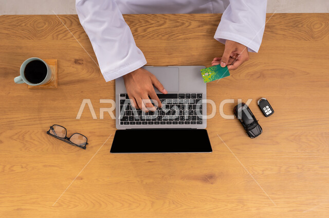 الدفع من خلال بطاقة الصراف البنكية، استخدام التكنولوجيا في طلب المستلزمات، عرض شاشة سوداء فارغة، البيع والشراء من الانترنت، صورة علوية عامودية لرجل عربي خليجي سعودي يستخدم الحاسب المحمول وبجانبه مجموعة سيارات، الحصول على السيارات الحديثة اونلاين