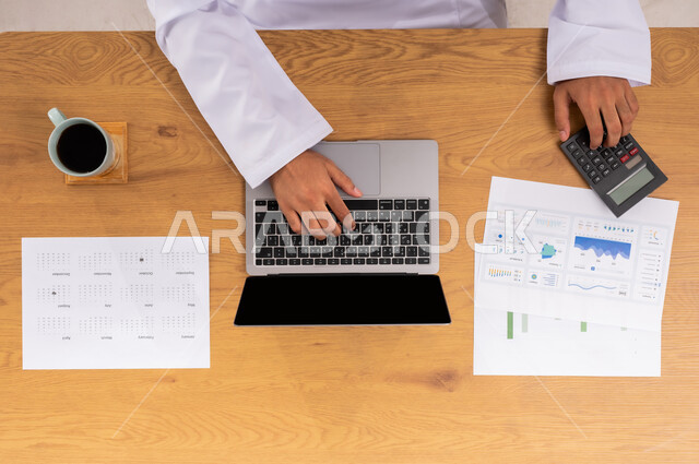 دمج الأجهزة الالكترونية بالعمل، عرض شاشة سوداء فارغة، الاسثمار المالي الالكتروني، متابعة مخطط الرسوم البيانية للنمو الاقتصادي، صورة عامودية علوية ليدي رجل عربي خليجي سعودي يقوم بدراسة وتحليل الاحصائيات عبر الحاسب المحمول، مراقبة الاقتصاد والمعلومات المالية