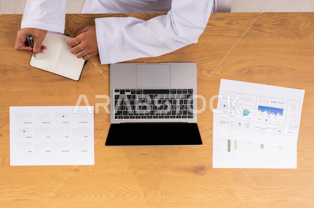 متابعة مخطط الرسوم البيانية للنمو الاقتصادي، صورة عامودية علوية ليدي رجل عربي خليجي سعودي يقوم بدراسة وتحليل الاحصائيات عبر الحاسب المحمول، مراقبة الاقتصاد والمعلومات المالية، دمج الأجهزة الالكترونية بالعمل، عرض شاشة سوداء فارغة، الاسثمار المالي الالكتروني