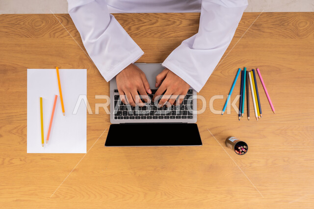 تعلم الأساليب الفنية عبر الانترنت، ممارسة موهبة الرسم والتلوين، الاهتمام بالنشاطات والهوايات، صورة علوية عامودية ليد رجل عربي  خليجي سعودي يستخدم الحاسب المحمول وبجانبه الأوراق والأقلام، الحصول على دفتر الرسم والألوان الخشبية أونلاين، عرض شاشة سوداء فارغة