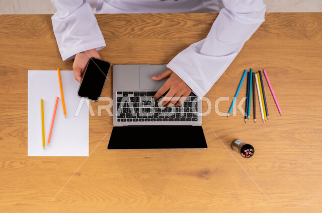 ممارسة موهبة الرسم والتلوين، الاهتمام بالنشاطات والهوايات، صورة علوية عامودية ليد رجل عربي  خليجي سعودي يستخدم اللابتوب والجوال بجانبه الأوراق والأقلام، الحصول على دفتر الرسم والألوان الخشبية أونلاين، عرض شاشة سوداء فارغة، تعلم الأساليب الفنية عبر الانترنت