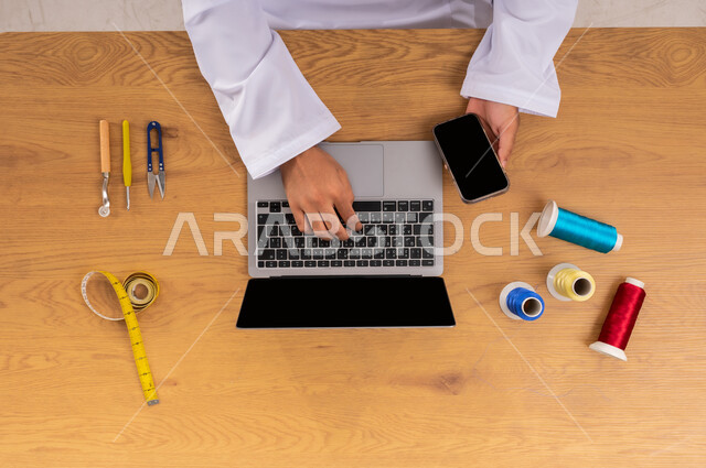 التعلم والتدريب من خلال المواقع الإلكترونية، عرض شاشة سوداء فارغة، صورة علوية عامودية ليدين رجل عربي خليجي سعودي يعمل على جهاز الحاسب المحمول والجوال وبجانبه مستلزمات الخياطة، البيع والشراء عبر الانترنت، الحصول على الأقمشة وأدوات الخياطة أونلاين