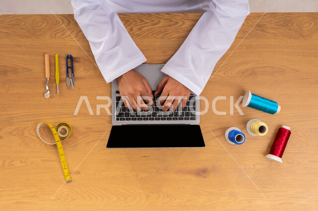 الحصول على الأقمشة وأدوات الخياطة أونلاين، التعلم والتدريب من خلال المواقع الإلكترونية، عرض شاشة سوداء فارغة، صورة علوية عامودية ليدين رجل عربي خليجي سعودي يعمل على جهاز الحاسب المحمول وبجانبه مستلزمات الخياطة، البيع والشراء عبر الانترنت