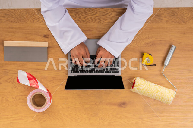 الاهتمام بالتجديد والتطور، الحصول على أدوات الصيانة والدهان عبر الإنترنت، سهولة شراء مستلزمات المهن أونلاين، صورة علوية عامودية ليد رجل عربي خليجي سعودي يقوم باستخدام الحاسب المحمول وبجانبه معدات التصليح، عرض شاشة سوداء فارغة