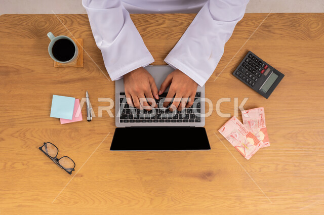 الأعمال والمهن المكتبية في السعودية, استخدام الآلة الحاسبة في تنظيم حسابات الشركة, صورة علوية عامودية ليدين رجل عربي سعودي خليجي التأكد من دفع الفواتير باستخدام جهاز الحاسب المحمول وبجانبه النقود، عرض شاشة سوداء فارغة، مراجعة العقود والإدارة الاقتصادية