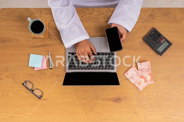 Contract review and economic management, office business and professions in Saudi Arabia, using calculator to organize company accounts, vertical top view of hands of Saudi Arabian Gulf man checking bills payment using laptop and mobile with cash next to him, blank black screen display