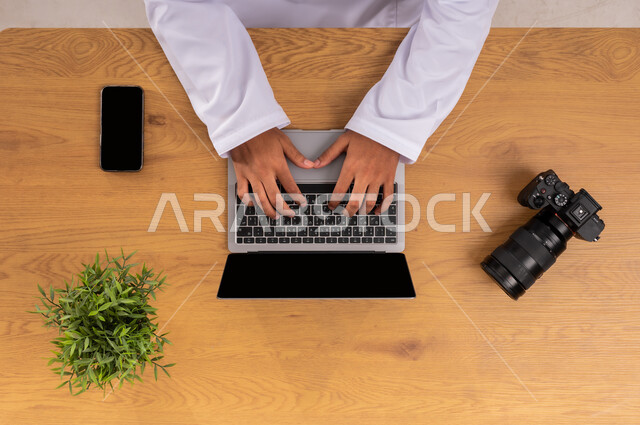 Mastering and professionalizing the profession of photography, learning to take artistic souvenir shots, practicing your favorite hobby with passion, displaying a blank black screen, a vertical overhead image of the hand of a Saudi Arabian Gulf man working on a mobile phone and next to him a photographic camera, using modern advanced technologies