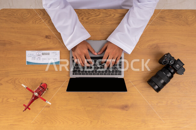 عرض شاشة سوداء فارغة، صورة علوية عامودية ليد رجل عربي خليجي سعودي يقوم بقطع تذاكر طيران عبر جهاز اللابتوب وبجانبه مجسم طائرة صغير وكاميرا فوتوغرافية، مفهوم السياحة والقيام بالجولات الترفيهية، الاستعداد لقضاء رحلة صيفية ممتعة، ممارسة هواية التصوير المفضلة