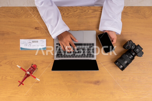 ممارسة هواية التصوير المفضلة، عرض شاشة سوداء فارغة، صورة علوية عامودية ليد رجل عربي خليجي سعودي يقوم بقطع تذاكر طيران عبر اللابتوب والجوال وبجانبه مجسم طائرة صغير وكاميرا فوتوغرافية، مفهوم السياحة والقيام بالجولات الترفيهية، الاستعداد لقضاء رحلة صيفية ممتعة