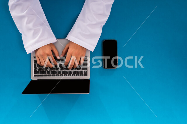 دمج العمل مع التقنية والتكنولوجيا الحديثة، مفهوم ادارة الاعمال وتحقيق الأهداف أونلاين، إنجاز المهام المطلوبة عن بعد، بورتريه علوي عامودي ليدين رجل عربي خليجي سعودي يعمل على الحاسب المحمول والجوال، عرض شاشة سوداء فارغة، مهن ووظائف مكتبية سعودية، خلفية زرقاء