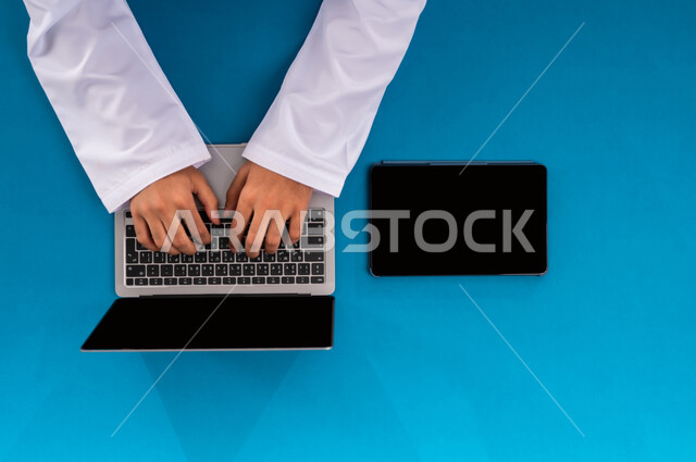 إنجاز المهام المطلوبة عن بعد، بورتريه علوي عامودي ليدين رجل عربي خليجي سعودي يعمل على الحاسب المحمول والتابليت، عرض شاشة سوداء فارغة، مهن ووظائف مكتبية سعودية، دمج العمل مع التقنية والتكنولوجيا الحديثة، مفهوم ادارة الاعمال وتحقيق الأهداف أونلاين، خلفية زرقاء