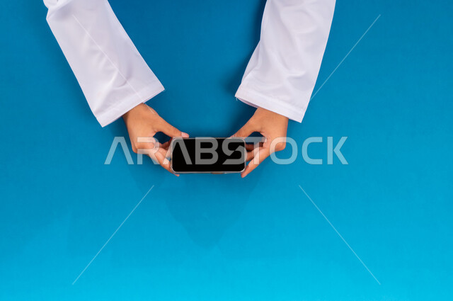 استخدام التقنيات الحديثة والمتطورة بالعمل، البيع والشراء عبر الانترنت، الاستفادة من تطبيقات التسوق أونلاين، عرض شاشة سوداء فارغة، إنجاز المهام المطلوبة عن بعد، بورتريه علوي عامودي ليدين رجل عربي خليجي سعودي يعمل على جهاز الهاتف المحمول، خلفية زرقاء