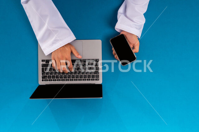 دمج العمل مع التقنية والتكنولوجيا الحديثة، مفهوم ادارة الاعمال وتحقيق الأهداف أونلاين، إنجاز المهام المطلوبة عن بعد، بورتريه علوي عامودي ليدين رجل عربي خليجي سعودي يعمل على الحاسب المحمول والجوال، عرض شاشة سوداء فارغة، مهن ووظائف مكتبية سعودية، خلفية زرقاء