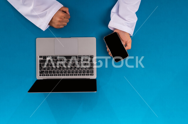 مهن ووظائف مكتبية سعودية، دمج العمل مع التقنية والتكنولوجيا الحديثة، مفهوم ادارة الاعمال وتحقيق الأهداف أونلاين، إنجاز المهام المطلوبة عن بعد، بورتريه علوي عامودي ليدين رجل عربي خليجي سعودي يعمل على الحاسب المحمول والجوال، عرض شاشة سوداء فارغة، خلفية زرقاء