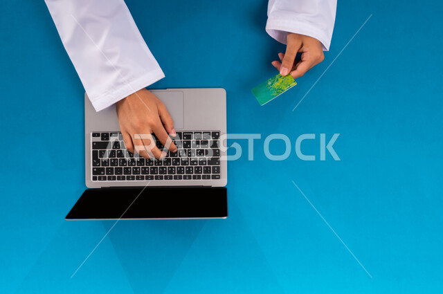 إدخال المعلومات البنكية على الحاسوب المحمول، الشراء والدفع عن بعد، بورتريه علوي عامودي ليدين رجل عربي سعودي خليجي  يقوم باستخدام بطاقة الإئتمان، التسوق الالكتروني عبر جهاز اللابتوب، إمكانية تسديد الفواتير بالبطاقة الالكترونية، عرض شاشة سوداء فارغة، خلفية زرقاء