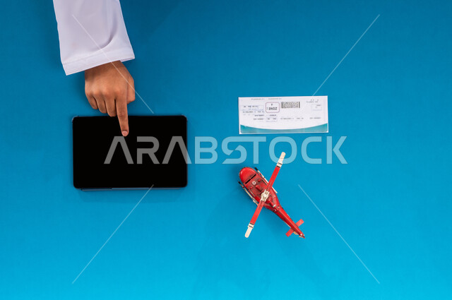 استخدام الانترنت للبحث عن رحلات جوية، مفهوم السياحة والقيام بالجولات الترفيهية، الاستعداد لقضاء رحلة صيفية ممتعة، عرض شاشة سوداء فارغة، بورتريه علوي عامودي ليد رجل عربي خليجي سعودي يستخدم جهاز التابليت وبجانبه مجسم طائرة، قطع تذاكر الطيران أونلاين، خلفية زرقاء