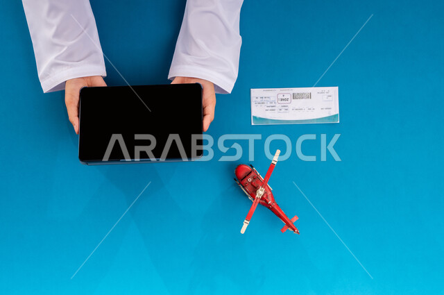 قطع تذاكر الطيران أونلاين، استخدام الانترنت للبحث عن رحلات جوية، مفهوم السياحة والقيام بالجولات الترفيهية، الاستعداد لقضاء رحلة صيفية ممتعة، عرض شاشة سوداء فارغة، بورتريه علوي عامودي ليد رجل عربي خليجي سعودي يمسك جهاز التابليت وبجانبه مجسم طائرة، خلفية زرقاء