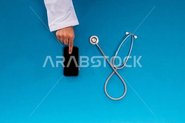 الخدمات والأجهزة الطبية، التسويق للأدوات الحديثة، بورتريه علوي عامودي ليدين رجل عربي سعودي خليجي يقوم بإجراء البحوث والدراسات عبر جهاز   الجوال وبجانب السماعة الطبية، عرض شاشة سوداء فارغة، طلب استشارة صحية أونلاين، مفهوم الطب والرعاية الصحية، خلفية زرقاء